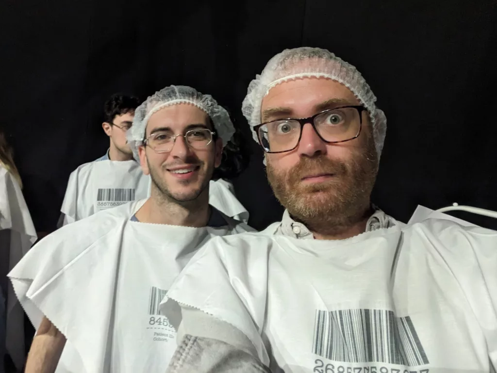 Two men wearing glasses, white hairnets, and white gowns with large barcodes printed on the chest, posing for a selfie against a dark background.