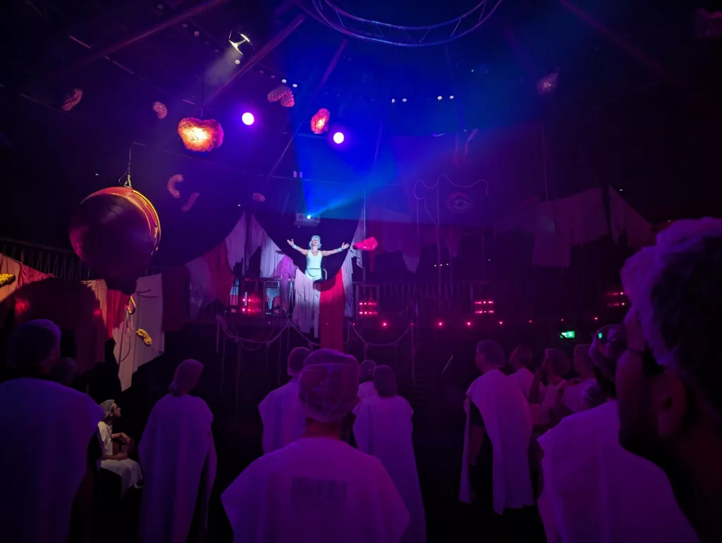 A performer with outstretched arms stands under a blue spotlight above an audience wearing white robes in a dark, atmospheric theatre space.