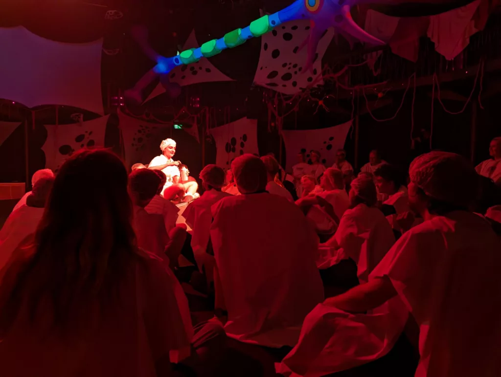 An audience in white smocks sits around a performer in a red-lit space under a glowing, colorful, neuron-like inflatable sculpture.