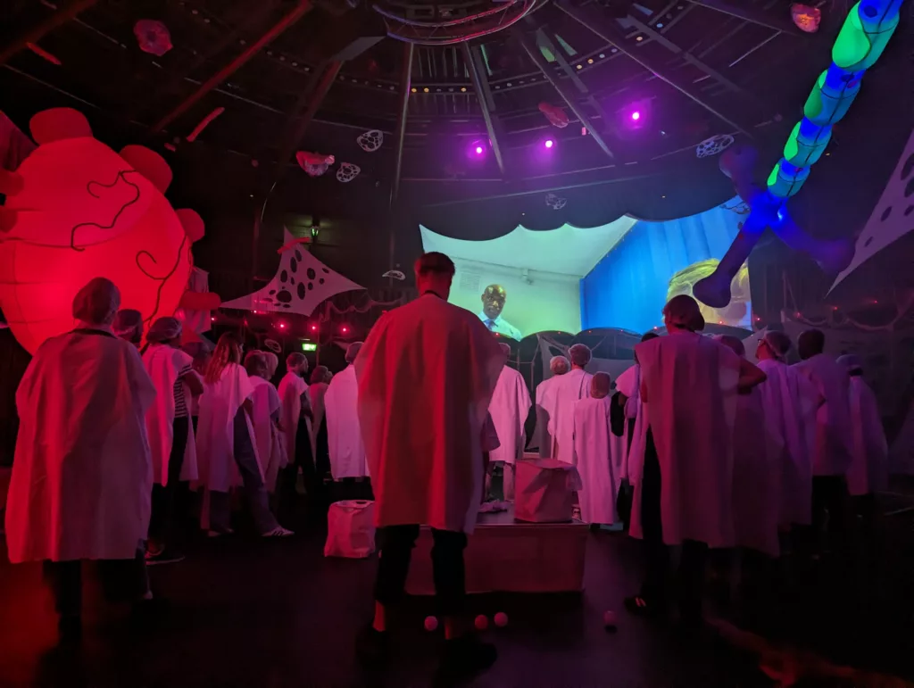 People in white capes watch a screen in a dark room with large glowing red and blue inflatable structures resembling body organs.