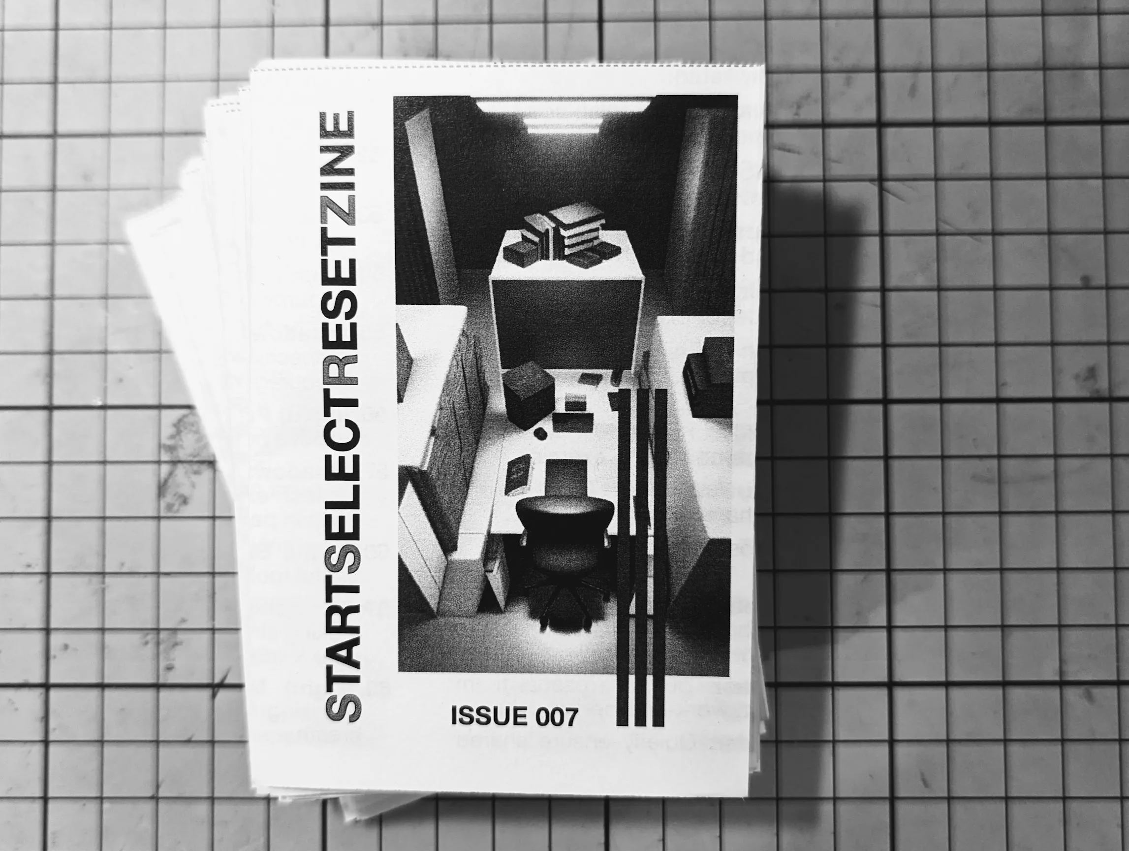 Black and white STARTSELECTRESETZINE Issue 007 on a gridded cutting mat. The cover features a grainy overhead illustration of a workspace.