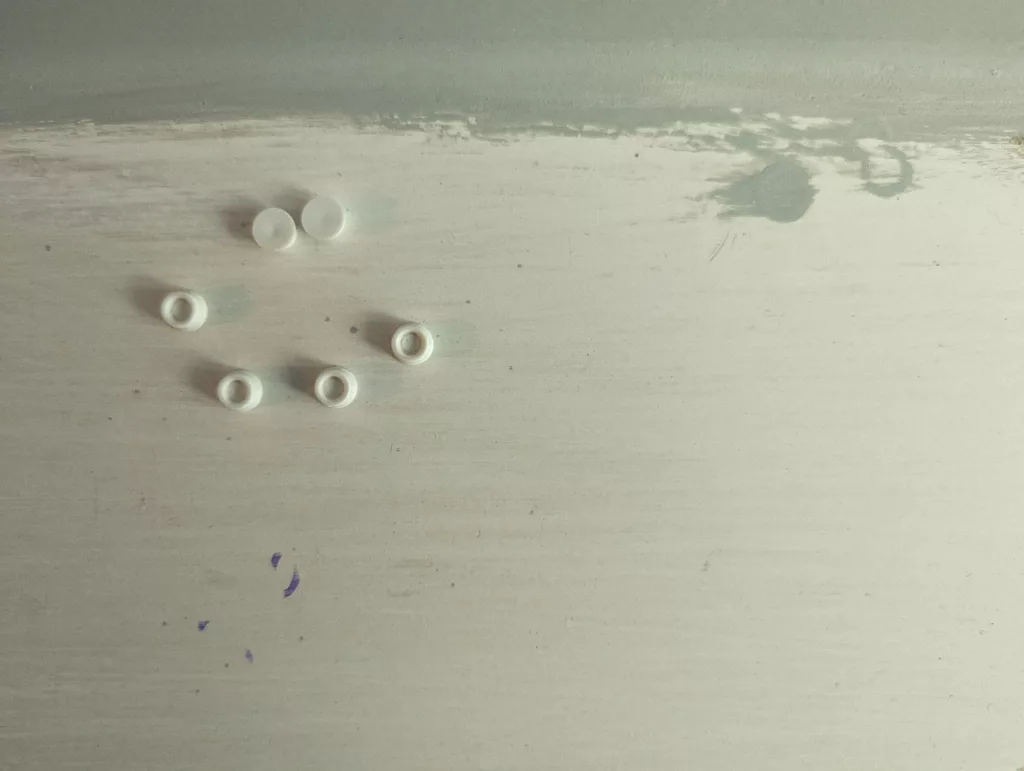 Six small white circular plastic pieces, including flat studs and rings, scattered on a worn white painted surface with purple ink marks.