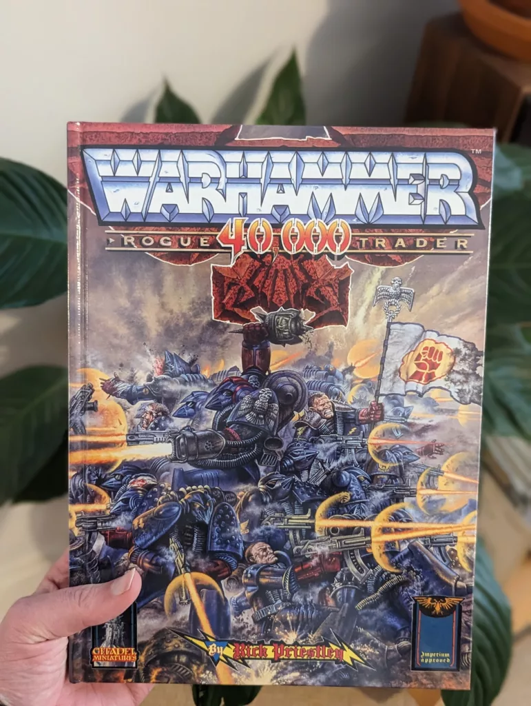 A hand holds the Warhammer 40,000 Rogue Trader rulebook by Rick Priestley, featuring retro sci-fi artwork of space marines in battle.