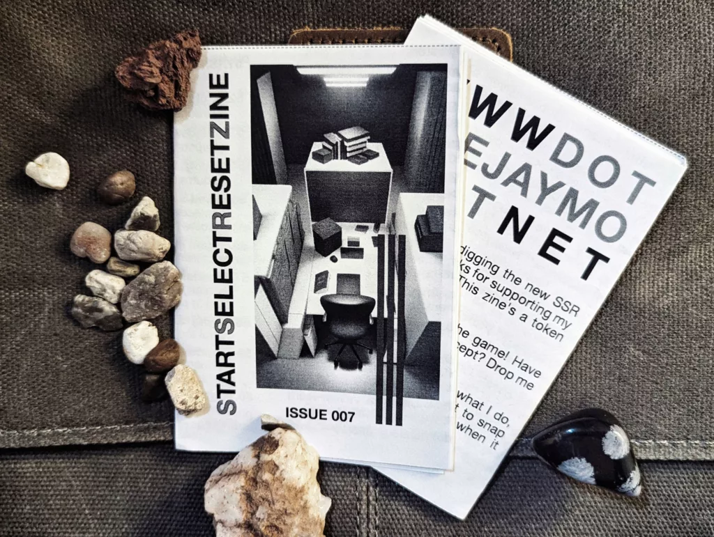 Featured image for STARTSELECTRESETZINE Issue 007 - The zine cover and a website card on grey fabric surrounded by small rocks.