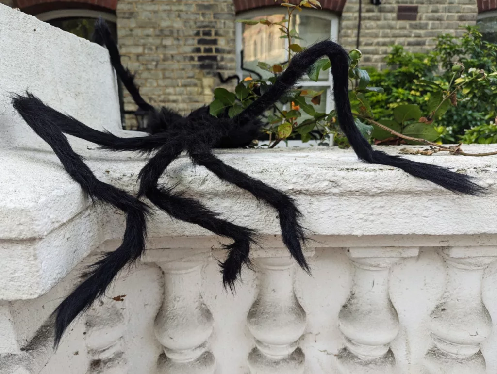 Large fuzzy black spider decoration with long spindly legs draped over a white stone balustrade in front of a brick building.
