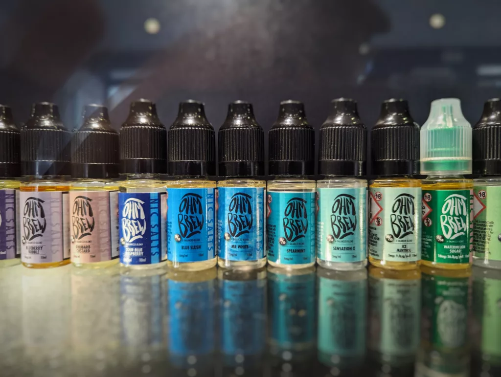 A row of colorful Ohm Brew nic salts e-liquid bottles reflecting on a dark counter, featuring flavors like Blue Slush and Ice Menthol.