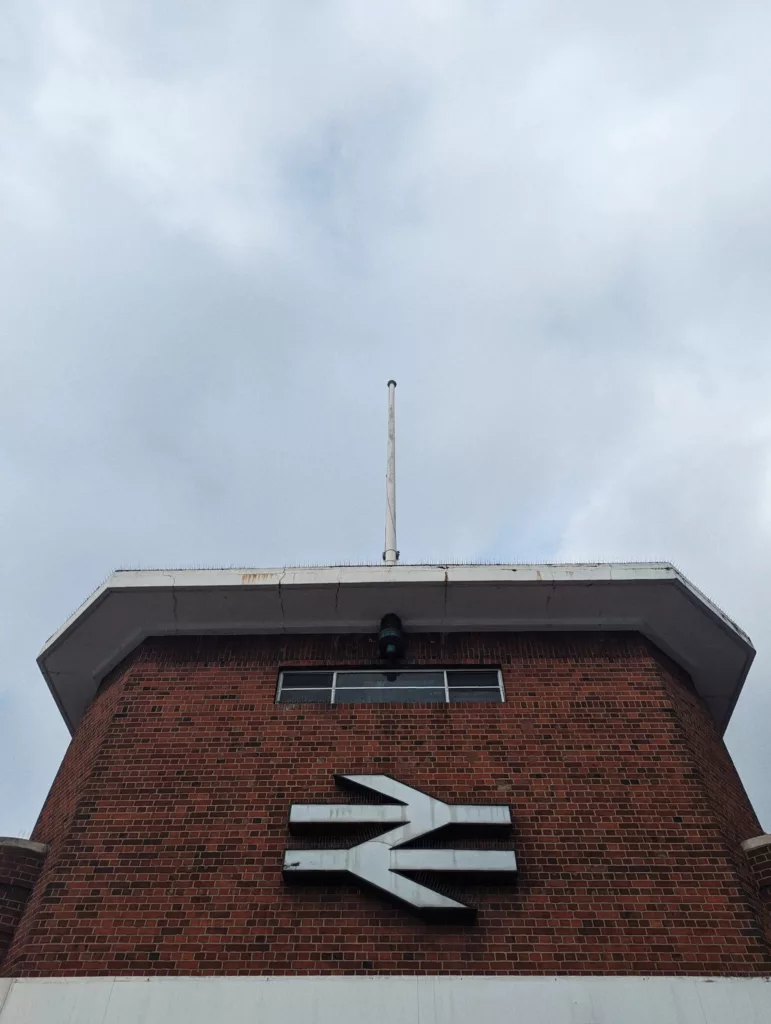 The white British Rail double arrow logo on a red brick station building with a flagpole on the roof under a grey overcast sky.