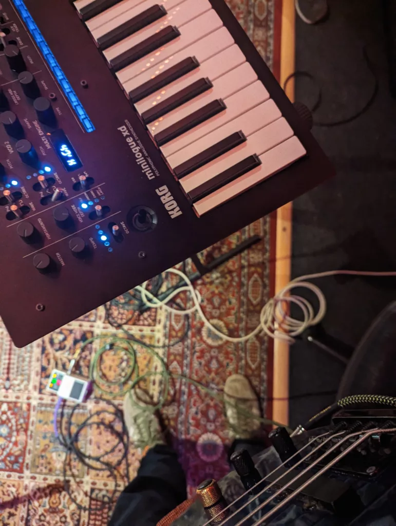 A Korg Minilogue xd synthesizer and guitar held above a patterned rug with scattered cables in a music studio.