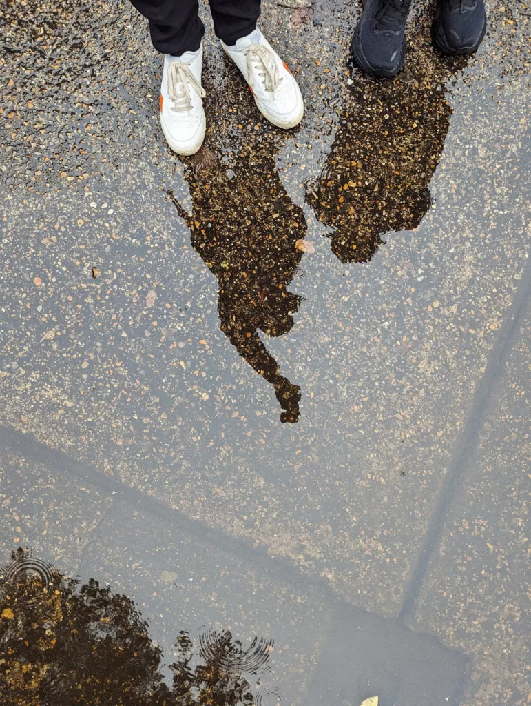 Two people's silhouettes reflected in a puddle on wet asphalt, with their white and black trainers visible at the top edge of the frame.