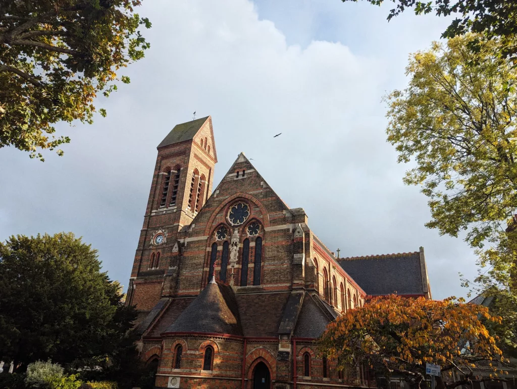 A red-brick gothic-style church with a tall tower and rose window, framed by yellow and green autumn trees under a cloudy blue sky.