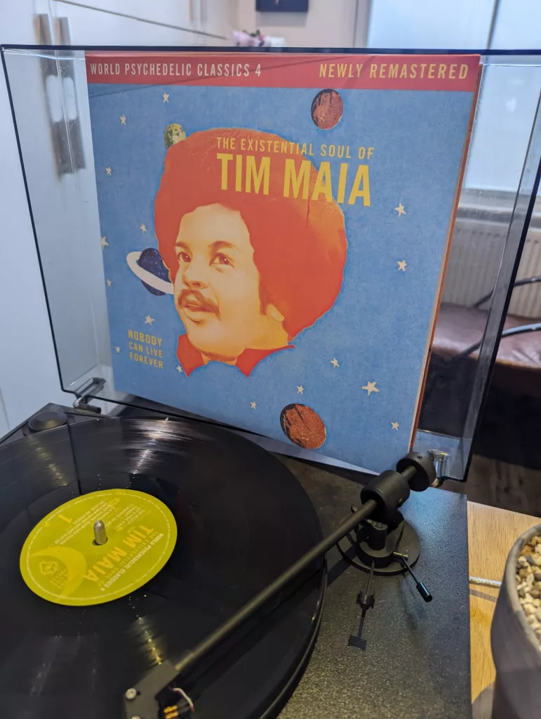 Tim Maia’s "The Existential Soul of Tim Maia" vinyl record plays on a black turntable with the blue, space-themed album cover displayed behind.
