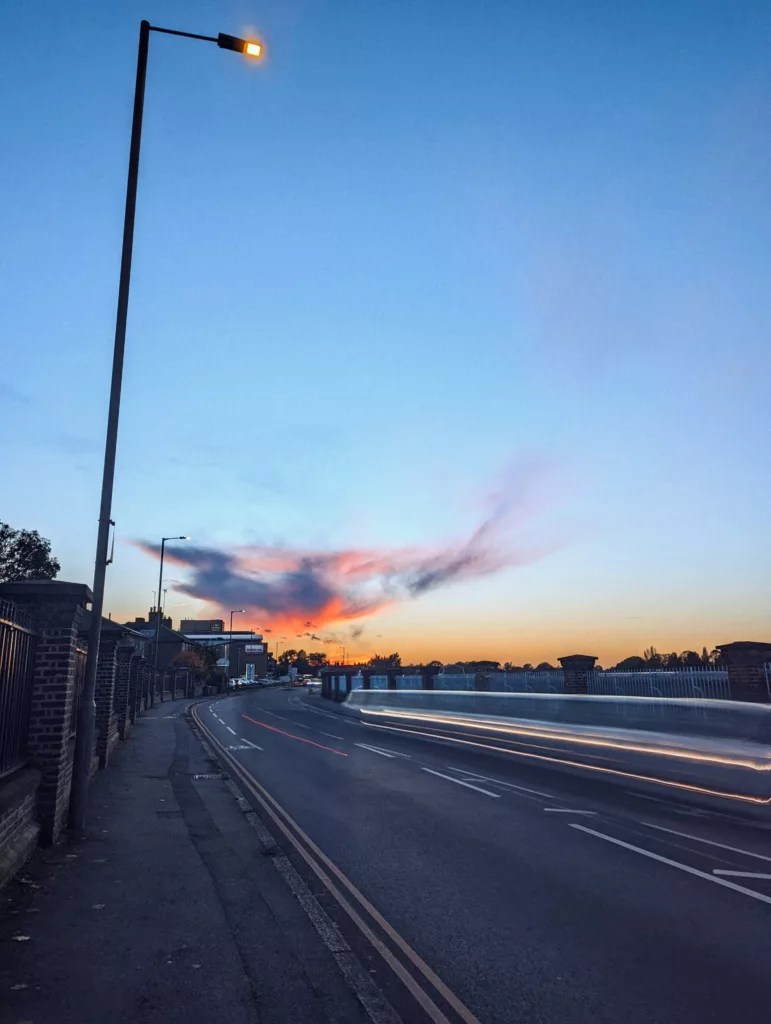 A long exposure of light trails from a vehicle on a road at dusk, beneath a tall streetlamp and a wispy, bird-shaped cloud lit by the sunset.