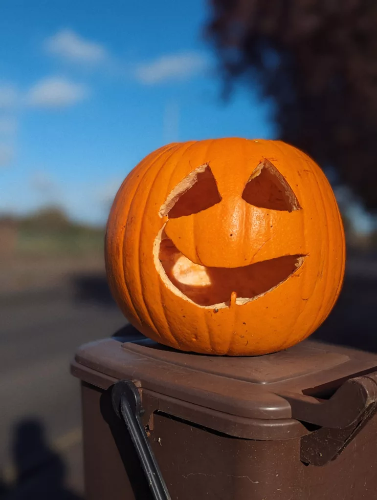 A smiling orange jack-o'-lantern sitting on a brown outdoor bin under a clear blue sky.