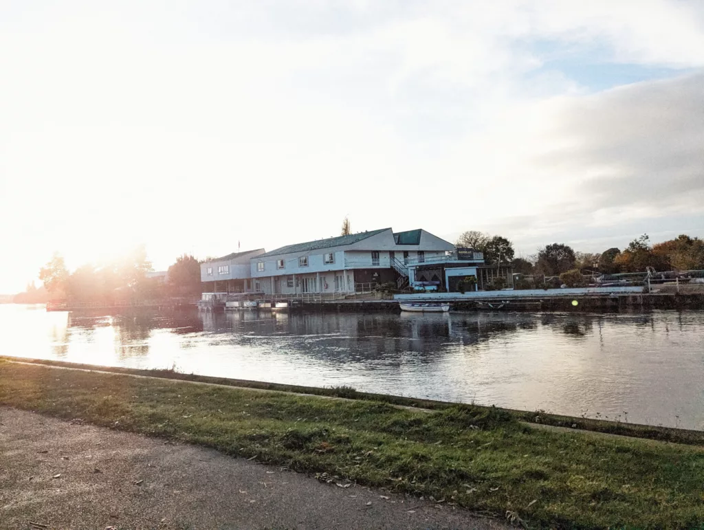 Sunlight flares over a wide river reflecting white buildings on the opposite bank, viewed from a grassy towpath.