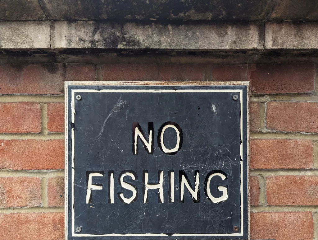 A weathered black sign with white lettering reading NO FISHING, screwed onto a red brick wall beneath a stone ledge.