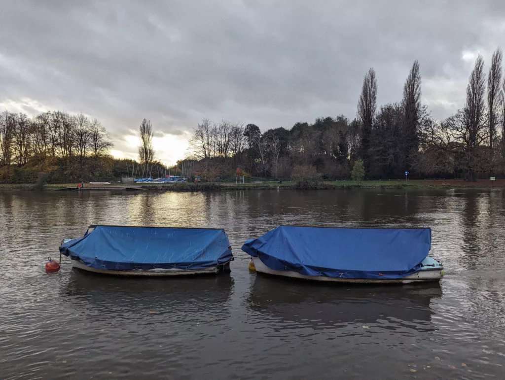Two boats with blue tarps moored on a calm river, backed by tall autumnal trees under a moody, overcast sky.