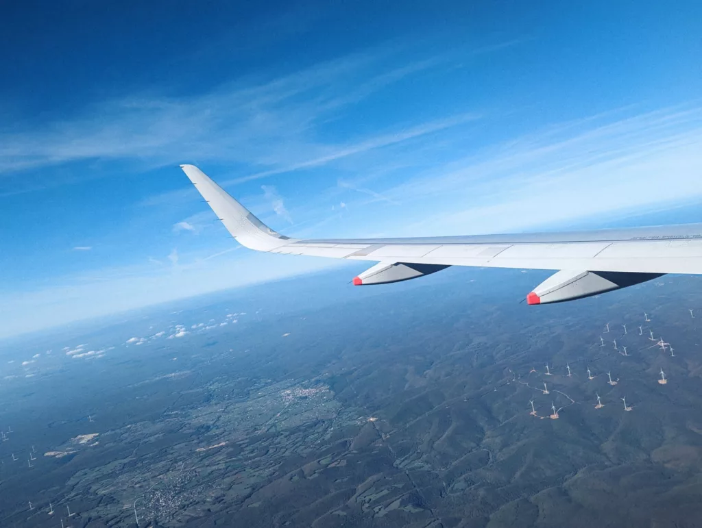 An airplane wing with a vertical winglet over a green hilly landscape dotted with numerous wind turbines under a bright blue sky.