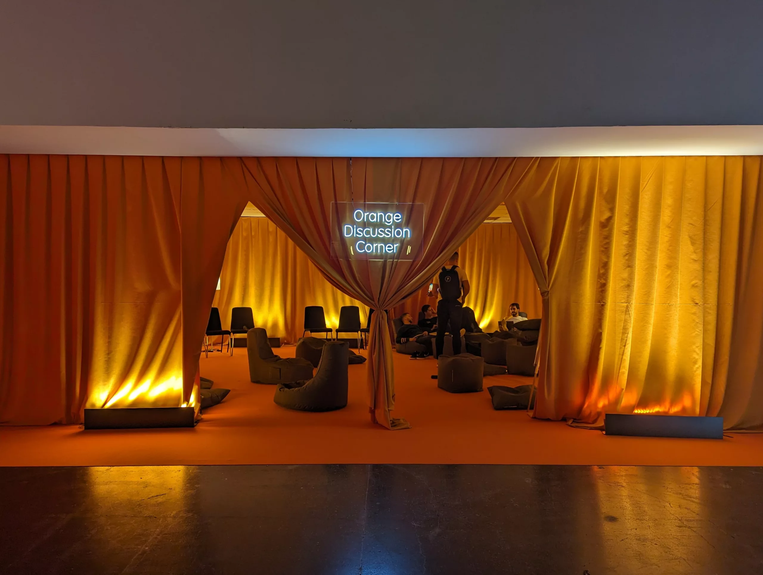 Orange Discussion Corner" neon sign above a room draped in orange curtains with bean bag chairs and warm floor lighting.