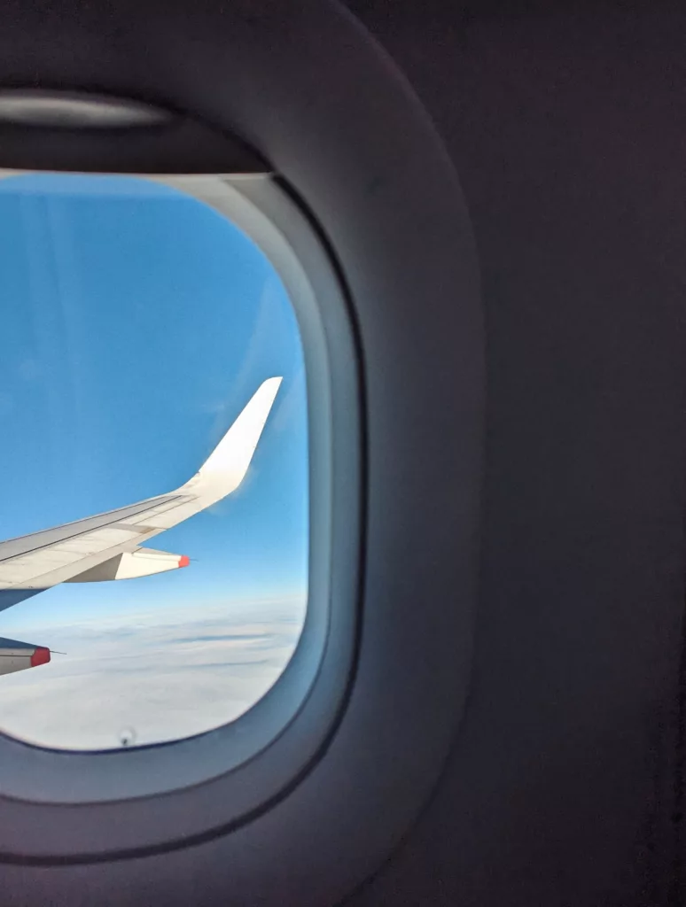 A white airplane wing with red tips seen through an oval cabin window against a bright blue sky and clouds below.