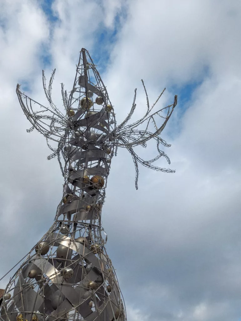 Spiralling wireframe holiday sculpture decorated with gold and silver baubles and fairy lights against a cloudy blue sky.