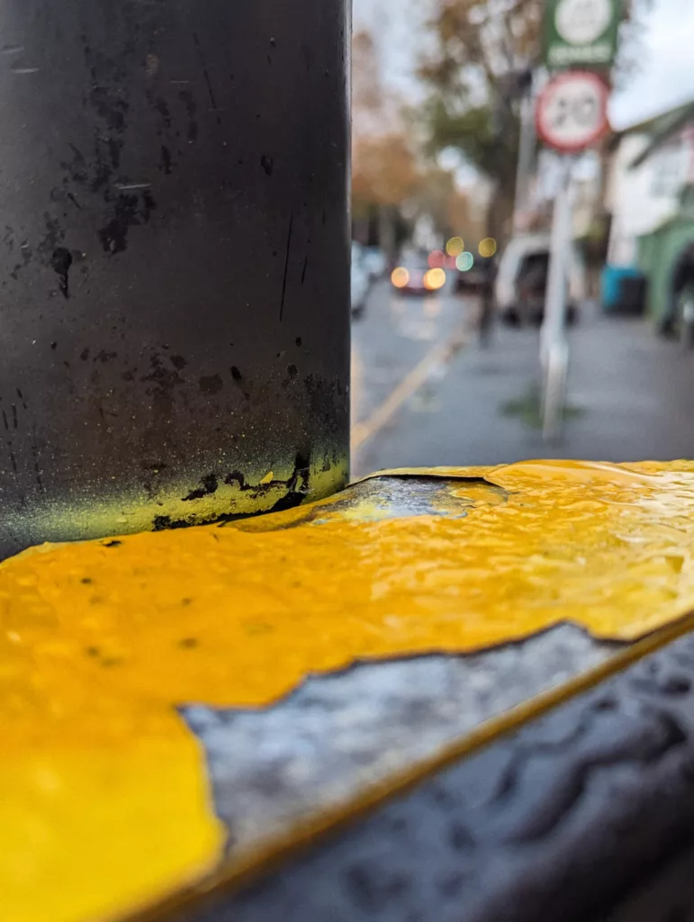 Close-up of rain-slicked peeling yellow paint on a metal bollard, with a blurred street scene and 20 mph sign in the background.