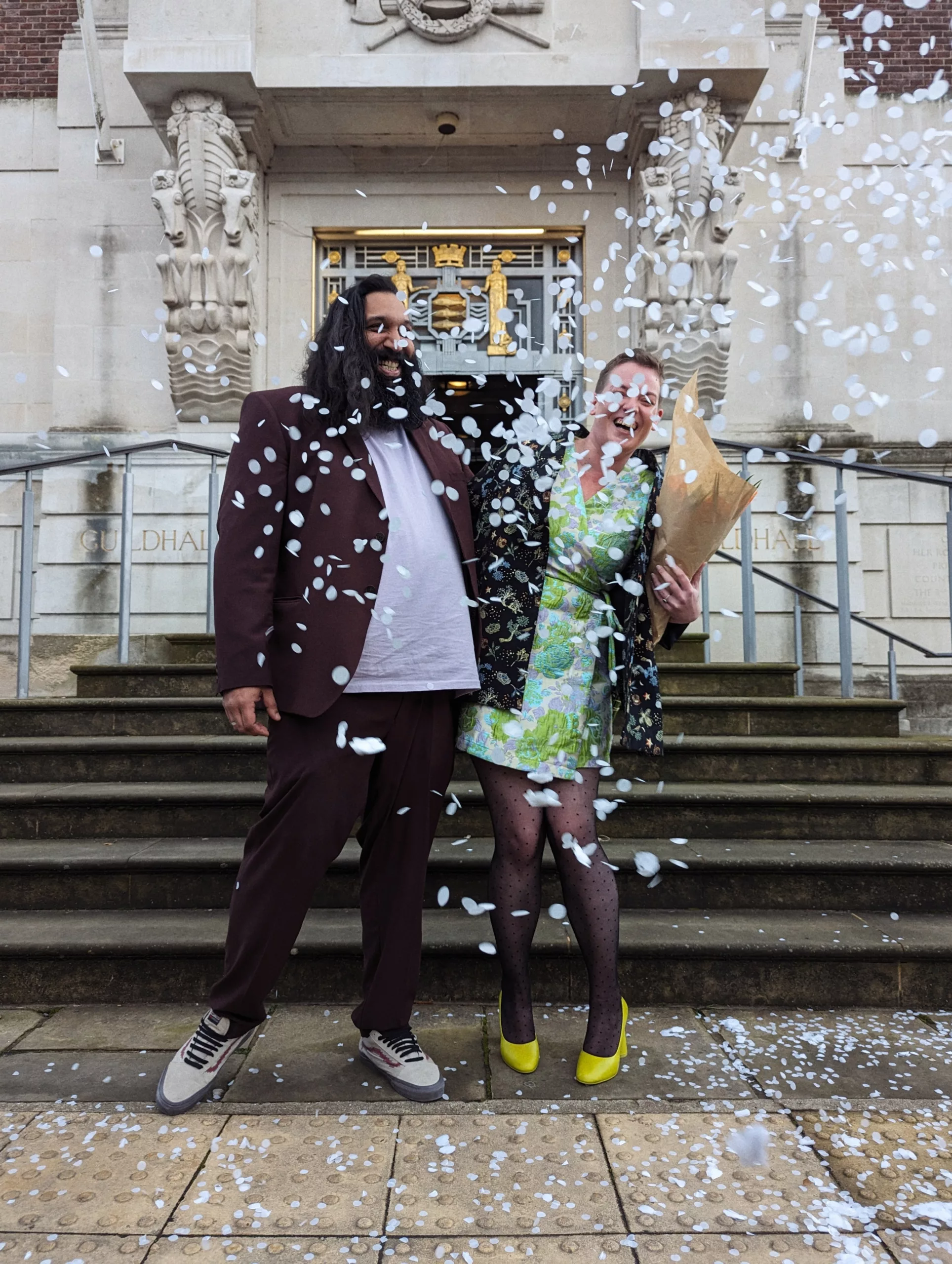 A laughing couple showered in white confetti on the stone steps of a Guildhall, dressed in a brown suit and a green floral dress.