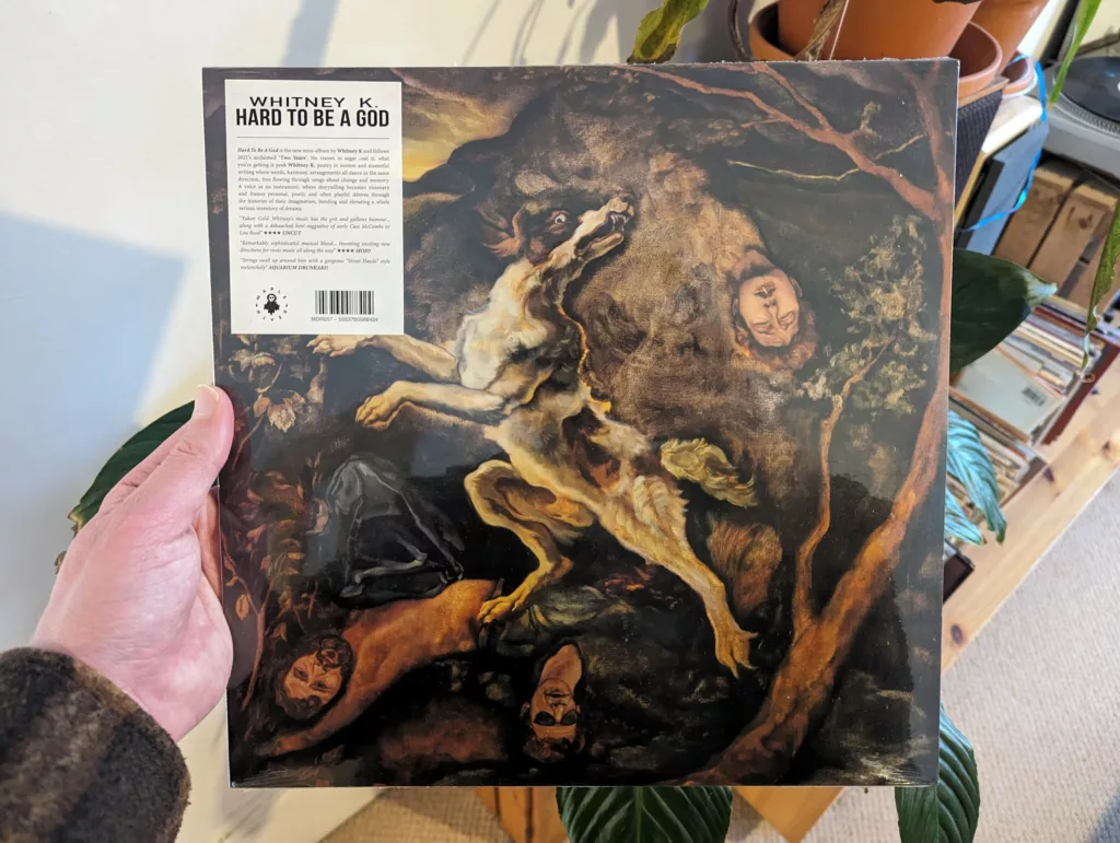 Hand holding Whitney K.'s Hard to be a God vinyl, featuring a painting of a dog leaping amongst human heads in a dark landscape.