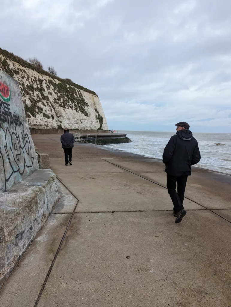 Two men walk along a concrete seaside promenade beneath towering white chalk cliffs under an overcast grey sky.