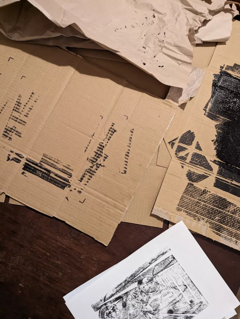 Brown cardboard sheets and crumpled paper with black ink smudges next to a white linocut print on a dark wooden surface.