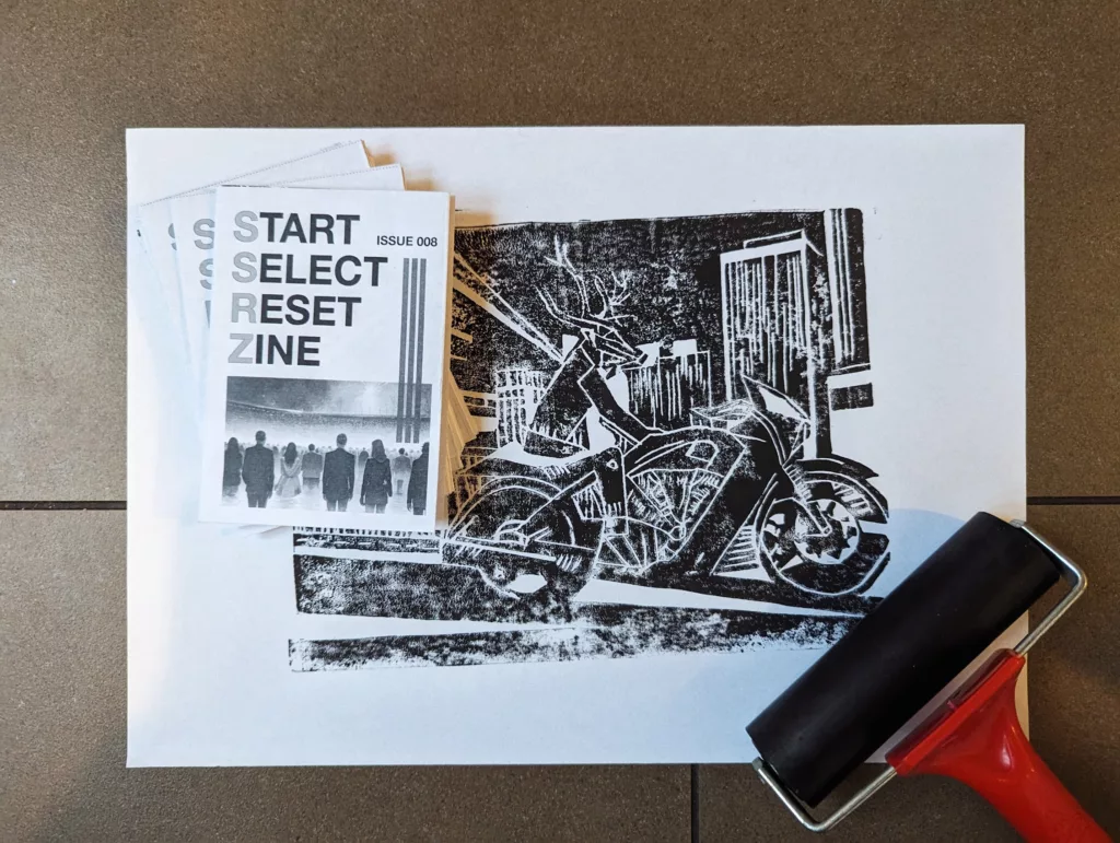 A stack of SSRZ Issue 008 zines next to a black and white linocut print of a deer riding a motorcycle, with a red ink brayer in the foreground.