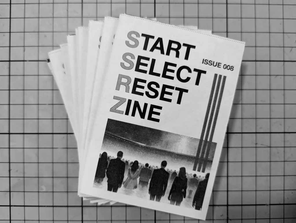 A stack of fanned-out black and white zines titled START SELECT RESET ZINE ISSUE 008 resting on a grid cutting mat.