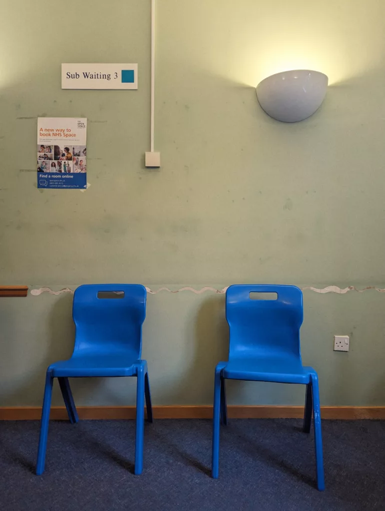 Two blue plastic chairs against a scuffed green wall in an NHS waiting area beneath a Sub Waiting 3 sign and a glowing wall lamp.