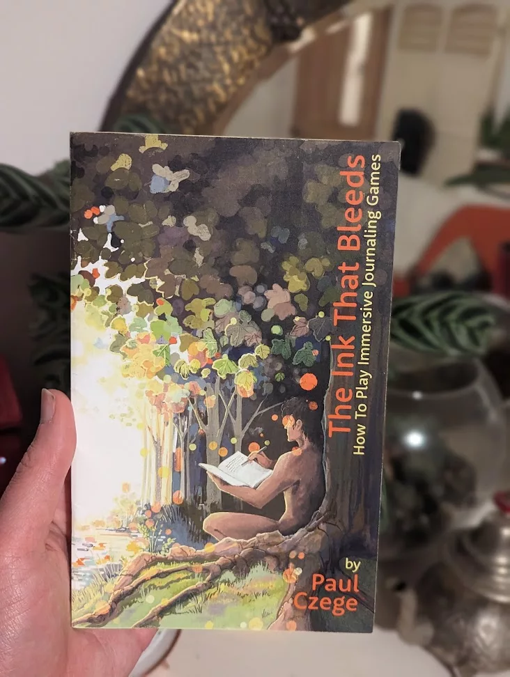Hand holding Paul Czege's book 'The Ink That Bleeds'. The cover art shows a person writing in a journal under a tree in a soft, glowing forest.
