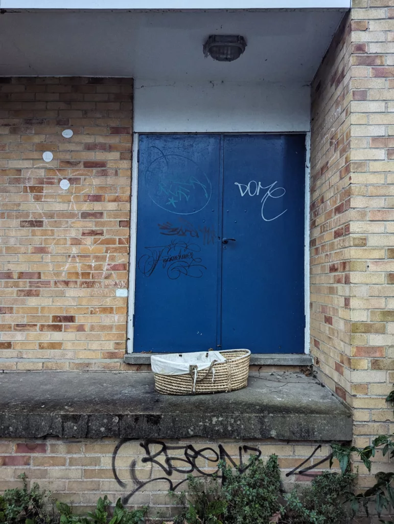 A woven Moses basket sits on a concrete ledge in front of blue double doors with graffiti tags, surrounded by yellow brick walls.