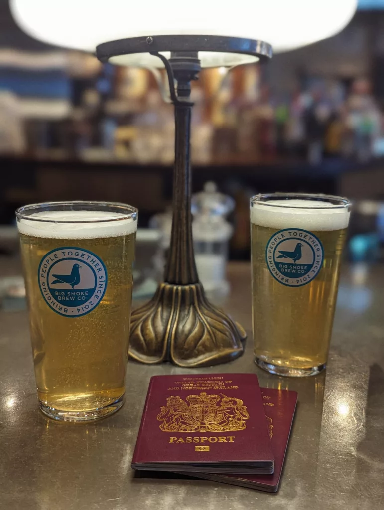Two pints of beer in Big Smoke Brew Co glasses and two British passports on a table under a decorative metal lamp in a pub.