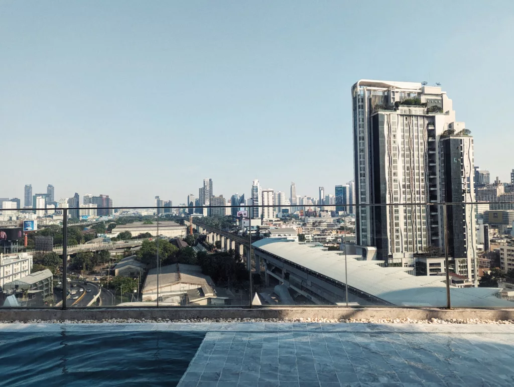 A rooftop pool overlooking a sprawling cityscape with modern skyscrapers and an elevated train station under a clear sky.