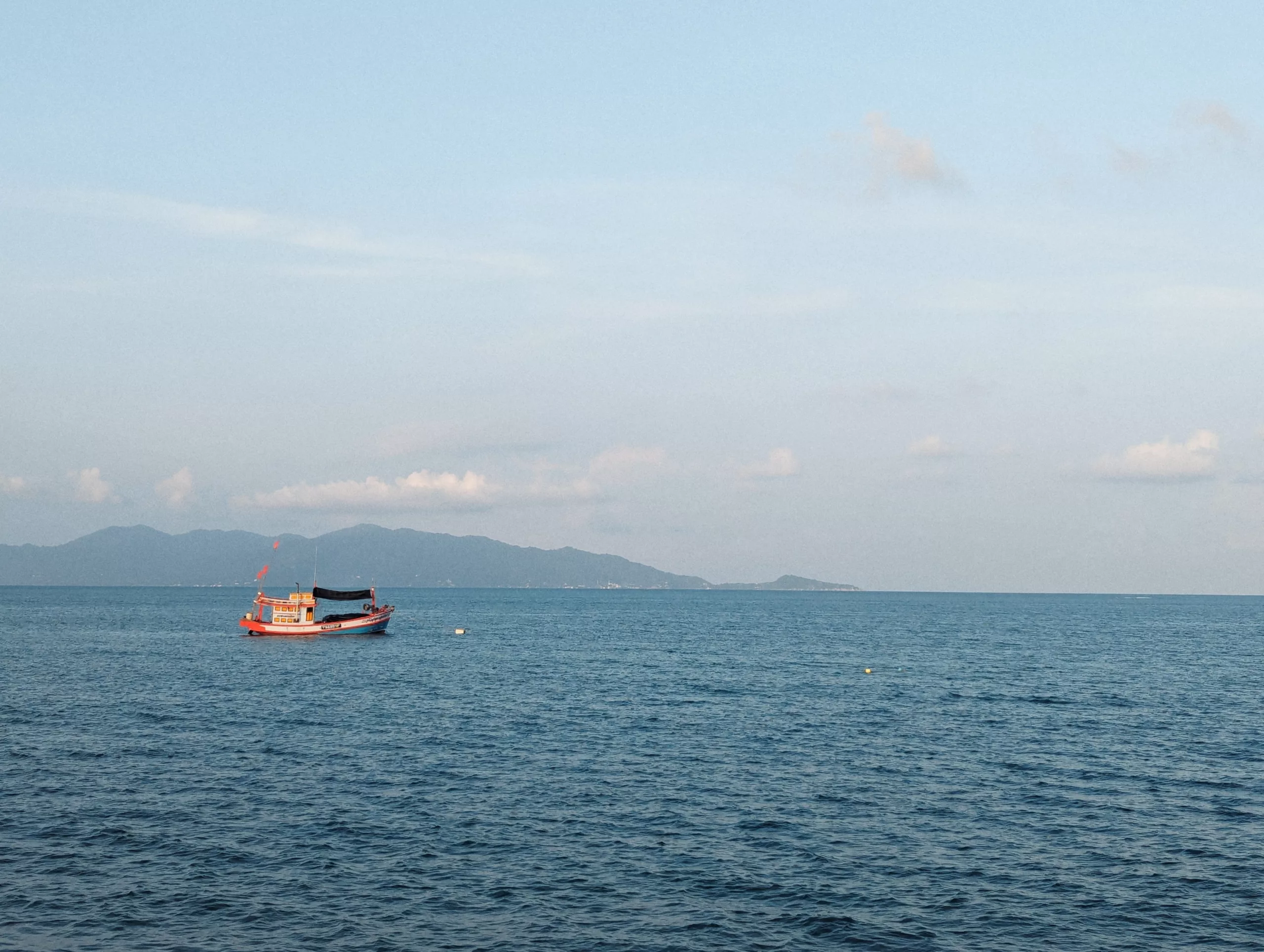 An orange and blue fishing boat floats on calm sea waters with rolling mountains in the distance under a pale, hazy sky.