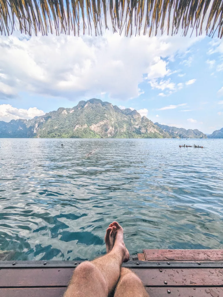 Bare legs resting on a wooden deck overlooking clear blue water and a large forested mountain, framed by a thatched roof at the top.