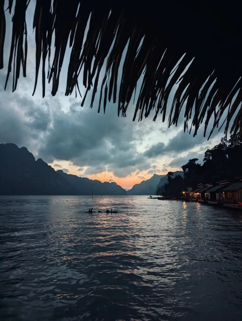 Floating lake houses glowing at twilight, framed by a thatched roof with mountain silhouettes against a cloudy orange sunset sky.