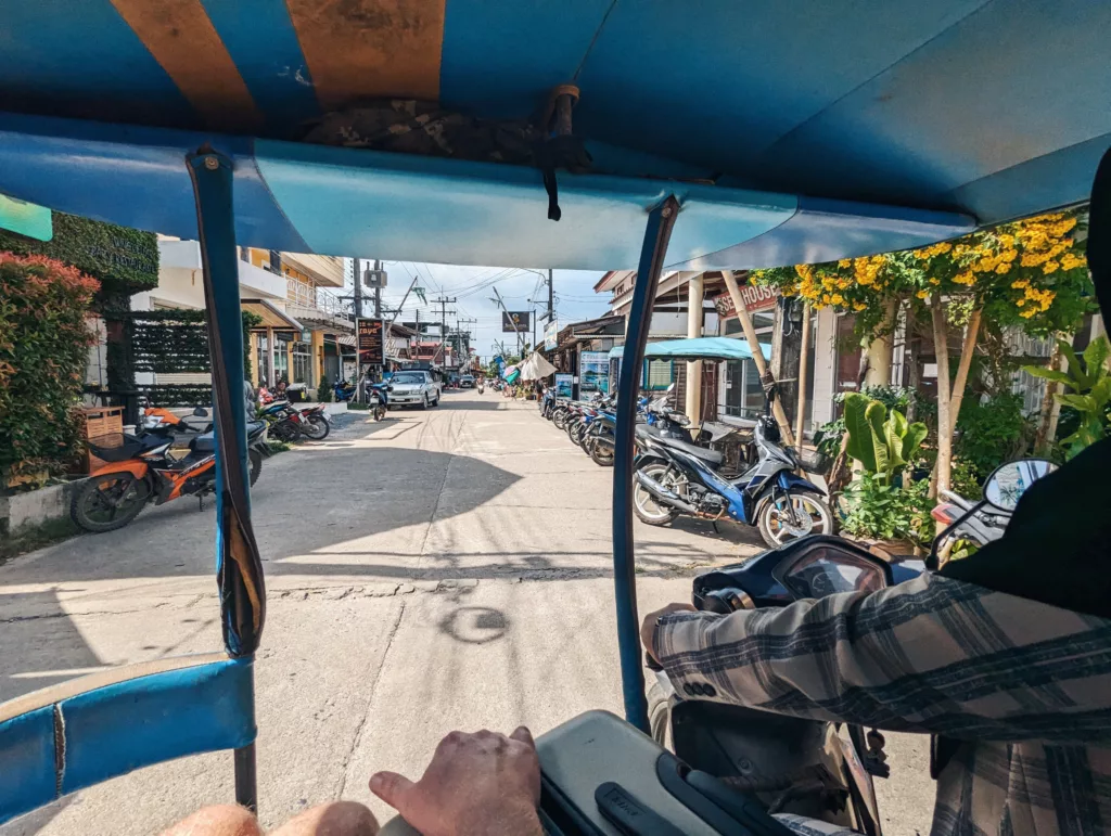 View from inside a blue tuk-tuk traveling down a sunlit street lined with parked motorbikes and small buildings.