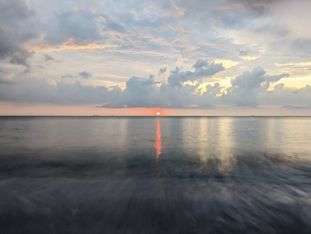 A long exposure of the sun setting over a calm ocean, casting a red reflection on the blurred water beneath a vast, cloudy sky.