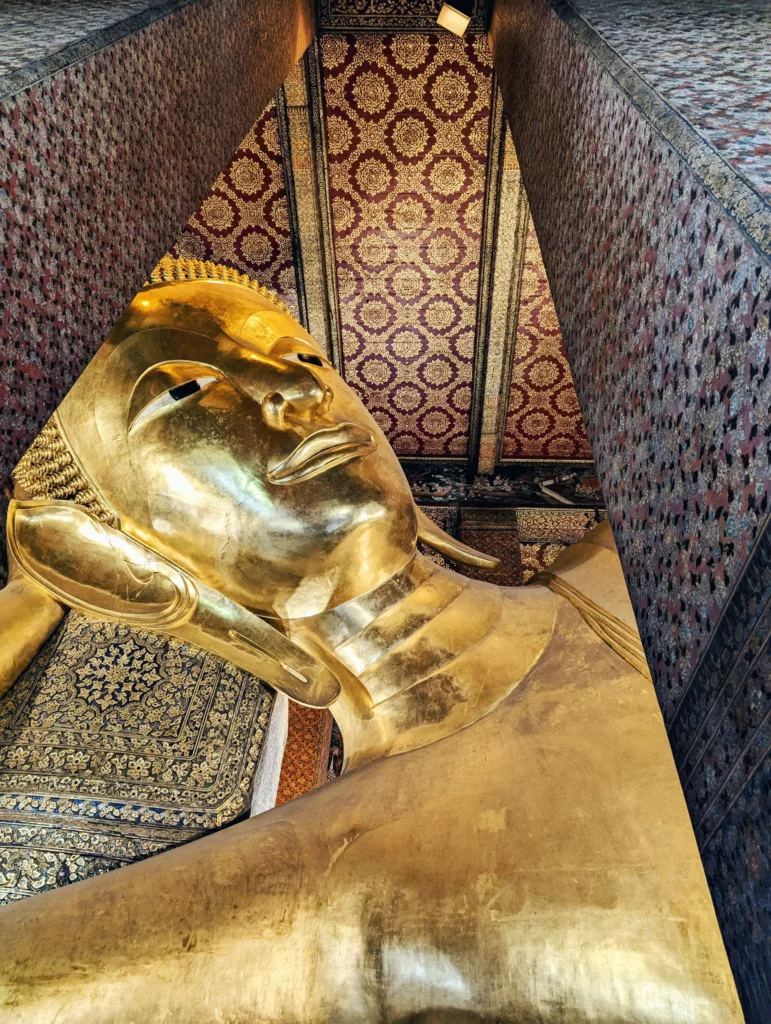 The gold face of the Reclining Buddha at Wat Pho, framed by patterned pillars and an ornate ceiling with red and gold floral motifs.