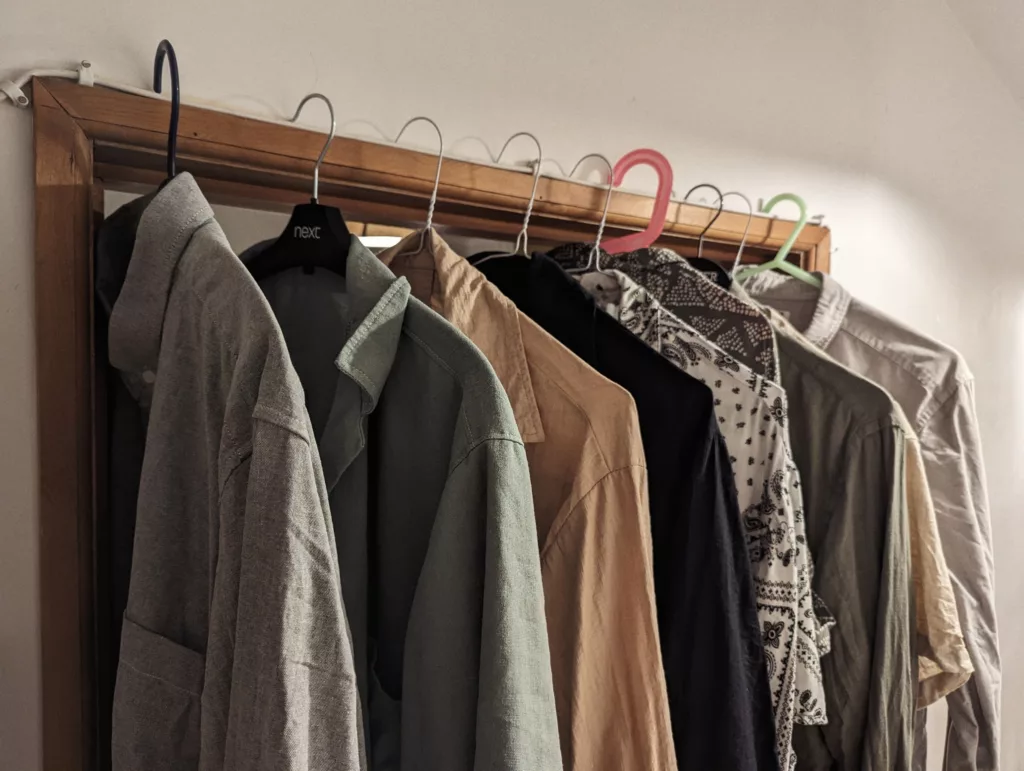 Shirts in shades of grey, tan, and green hanging from a row of mismatched hangers hooked over the top of a wooden door frame.