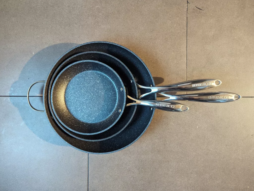 Three nested black speckled Neverstick2 frying pans with silver handles on a grey tiled surface.