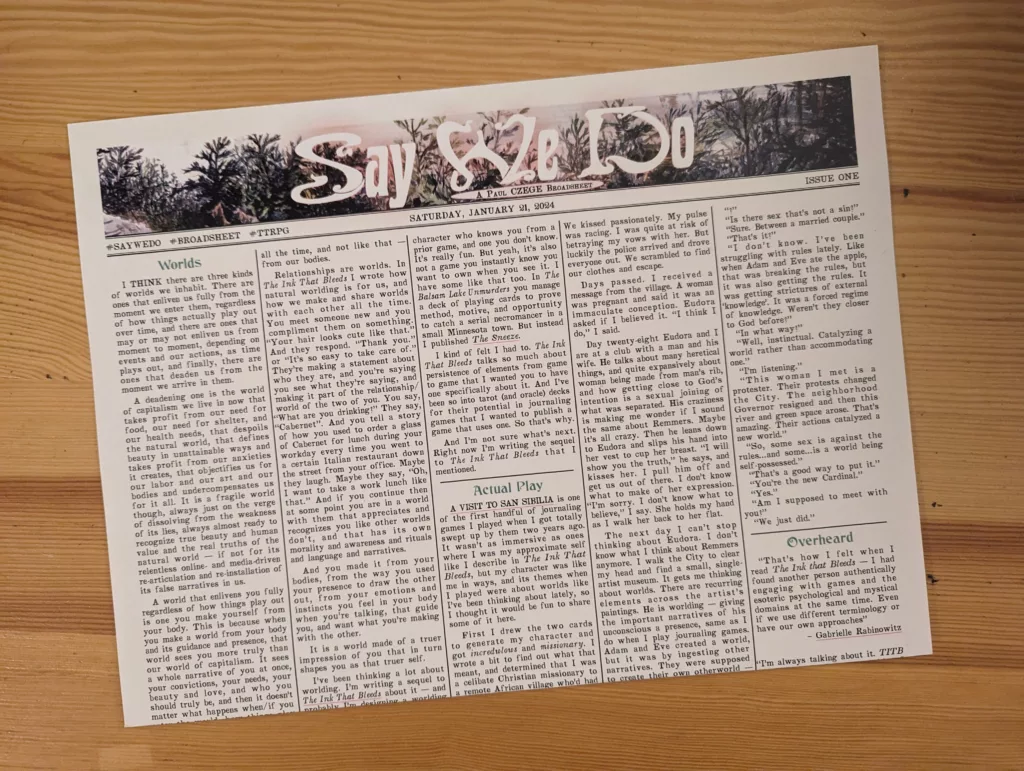 Say We Do broadsheet zine by Paul Czege, Issue One, featuring text columns and a forest-themed header, resting on a light wood surface.
