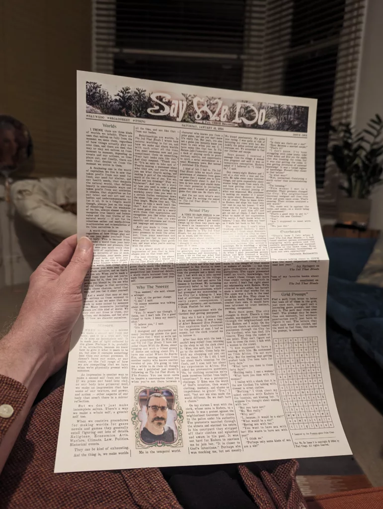 A hand holding a printed newsletter titled Say We Do, featuring dense columns of text and a small portrait of a man against a blurred background.