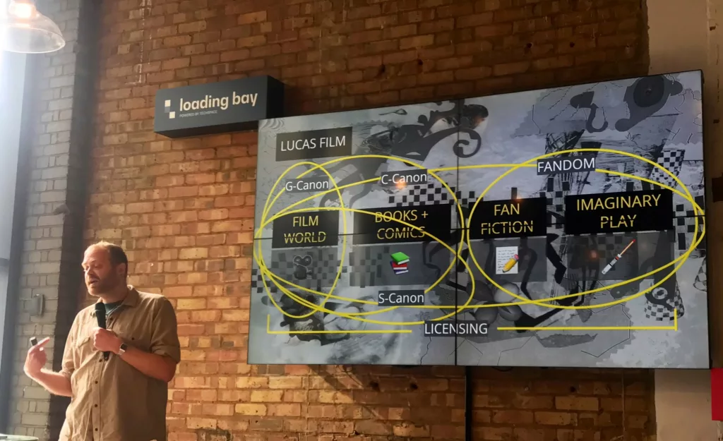 Featured image for Open Croissant - a man gives a presentation about Lucasfilm canon and fandom on a large screen in a brick-walled venue.
