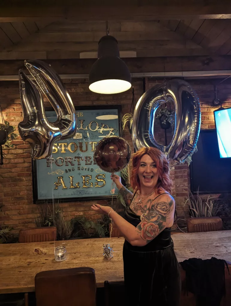 Smiling woman with red hair and tattoos celebrates her 40th birthday with large silver balloons in a rustic pub with a brick wall.