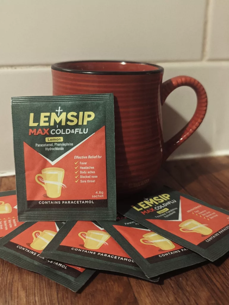 Lemsip Max Cold & Flu lemon sachets fanned out on a wooden surface in front of a red ceramic mug against a white tiled wall.