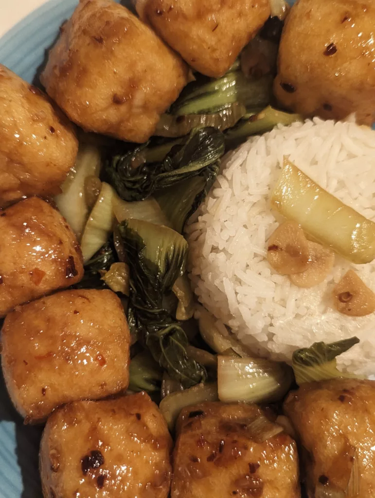 Golden glazed tofu cubes, sautéed bok choy, and a mound of white rice topped with garlic chips on a blue plate.